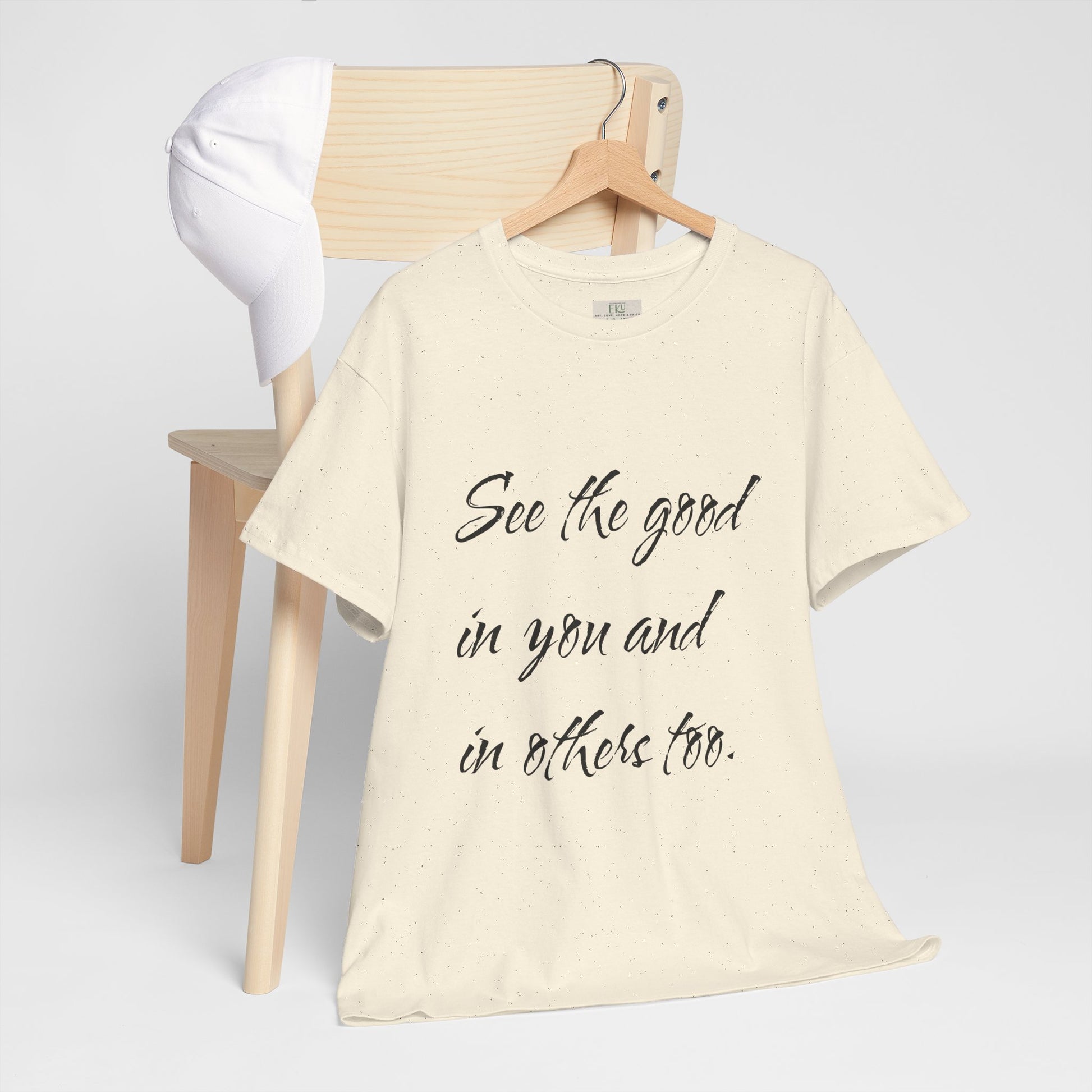 Natural/beige t-shirt with motivational quote text on a wooden hanger - hanging on wooden chair backing with a white cap on it.