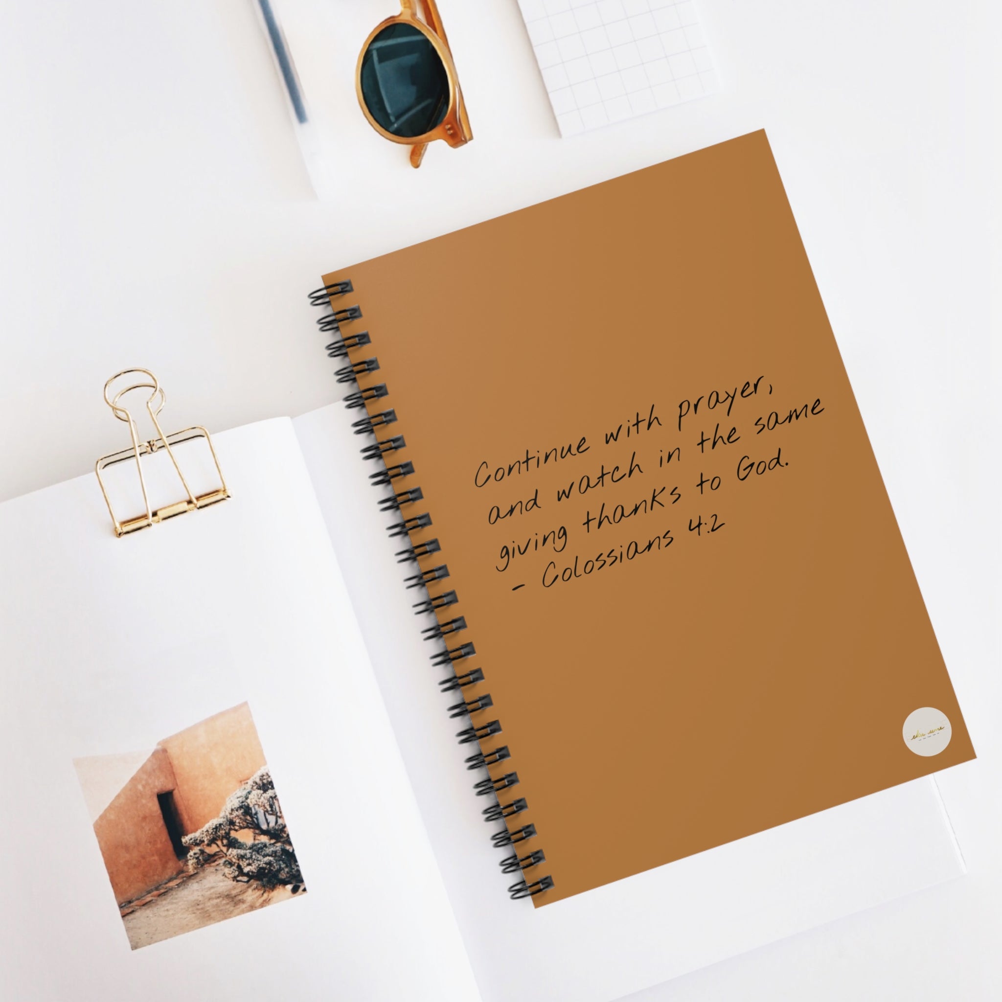 Spiral journal/notebook with 'Colossians 4:2' scripture text and eKuumeArt logo on the front cover, displayed on a light surface with a clip, photo and sunglasses