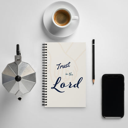 Spiral journal/notebook with 'Trust in the Lord' text on the cover – displayed on an off-white surface with a pencil, cup of coffee on a plate, tea pot and phone around it.