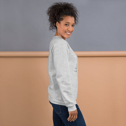 Woman wearing ash sweater with 'Believe' handwritten text and eKuumeArt logo on torso – angled view, featuring a grey and brown room setting.