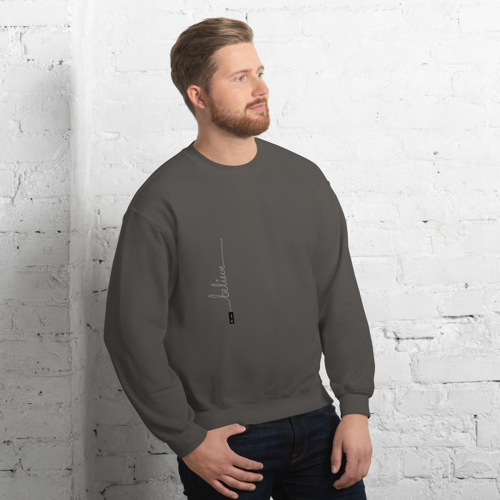 Man wearing charcoal sweater with 'Believe' handwritten text and eKuumeArt logo on torso – angled view, featuring a white brick wall room setting.