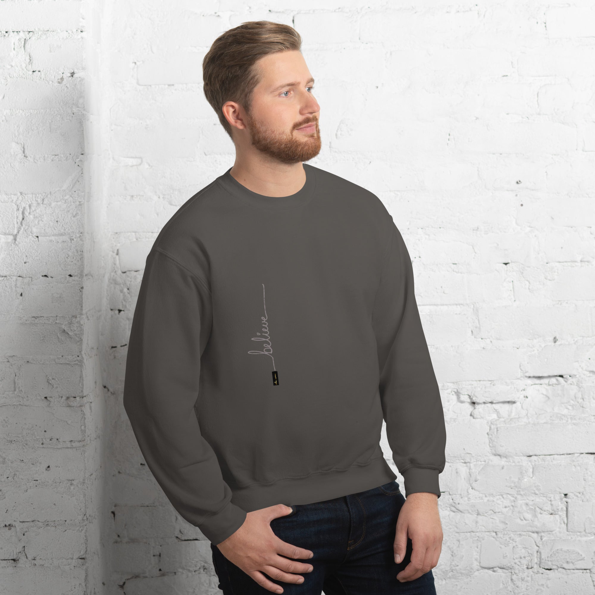 Man wearing charcoal sweater with 'Believe' handwritten text and eKuumeArt logo on torso – angled view, featuring a white brick wall room setting.