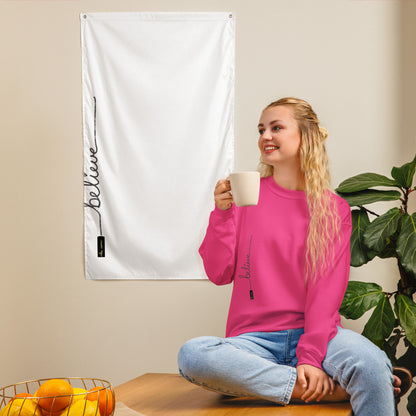 Woman wearing heliconia pink 'Believe' sweater – sitting on a wooden countertop holding a cup, near a plant and white tapestry with same text and logo on neutral wall.