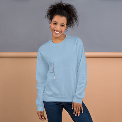 Woman wearing light blue sweater with 'Believe' handwritten text and eKuumeArt logo on torso – front view, featuring a grey and brown room setting.