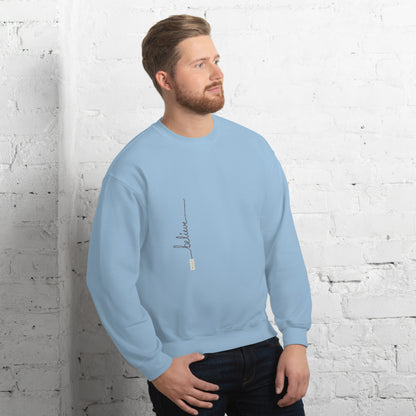 Man wearing light blue sweater with 'Believe' handwritten text and eKuumeArt logo on torso – angled view, featuring a white brick wall room setting.