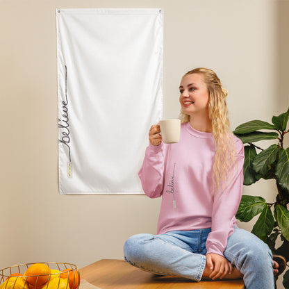 Woman wearing light pink 'Believe' sweater – sitting on a wooden countertop holding a cup, near a plant and white tapestry with same text and logo on neutral wall.