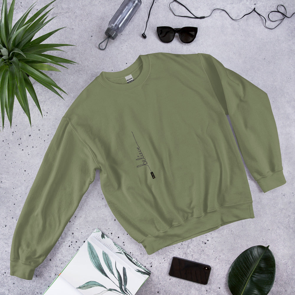 Military Green sweater with 'Believe' handwritten text and eKuumeArt logo on torso - featured on a stone surface with plant, journal and other daily items around it