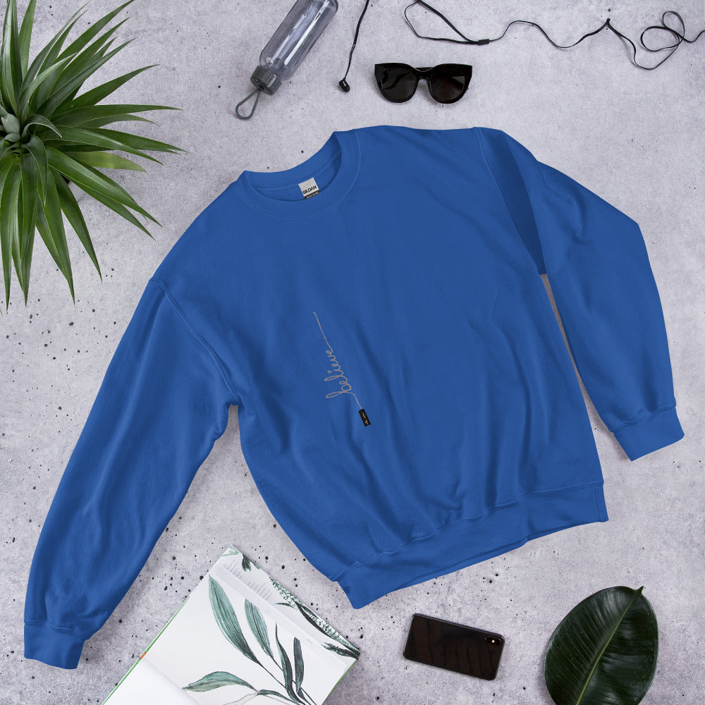 Royal sweater with 'Believe' handwritten text and eKuumeArt logo on torso – featured on a stone surface with plant, journal and other daily items around it.