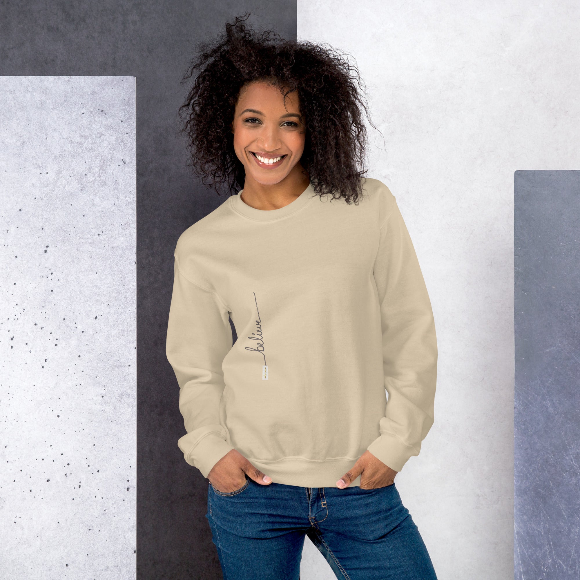 Woman wearing sand sweater with 'Believe' handwritten text and eKuumeArt logo on torso – front view, featuring shades of grey and white room setting.