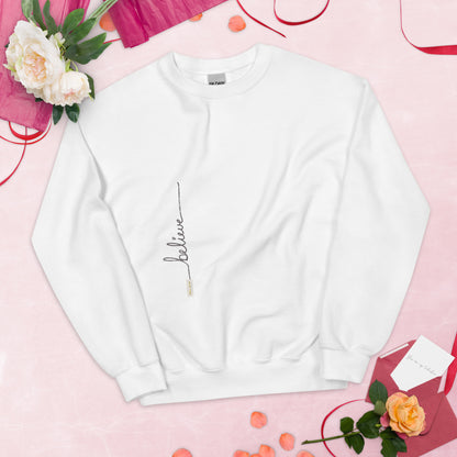 White sweater adorned with 'Believe' handwritten text and eKuumeArt logo on torso – displayed on light pink surface decorated with roses, petals and love cards.
